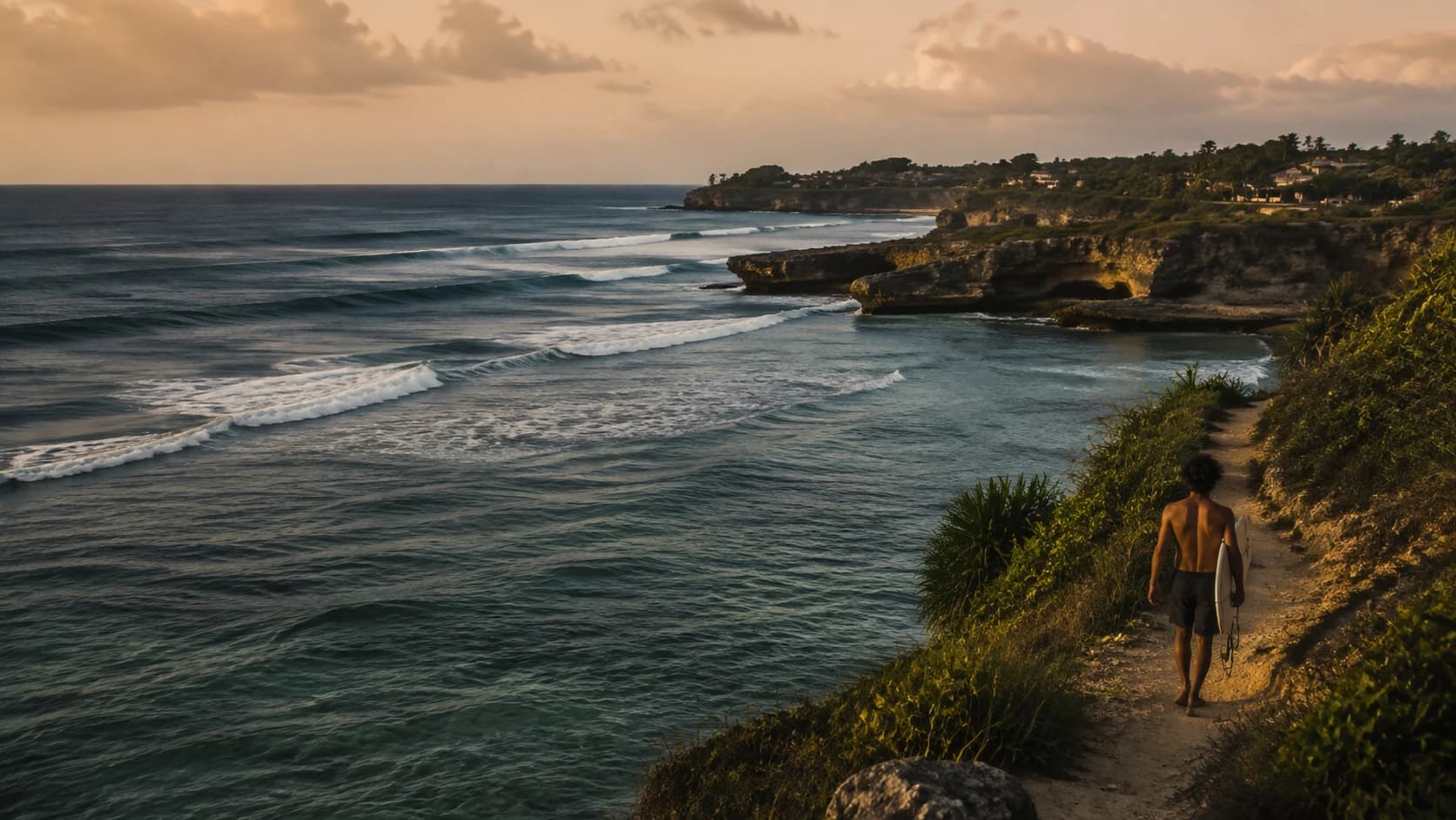 A surfer walking a coastal path above Indonesian reef waves at golden hour